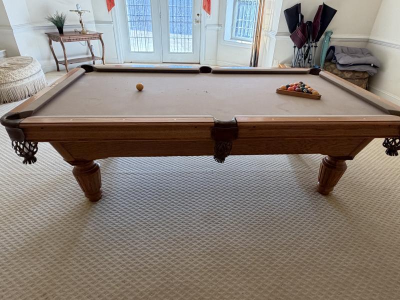Wooden pool table with cues and balls in a room with a white wall and window.