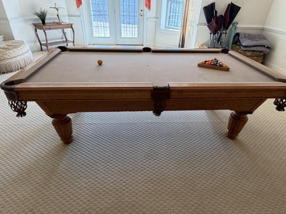 Wooden pool table with cues and balls in a room with a white wall and window.